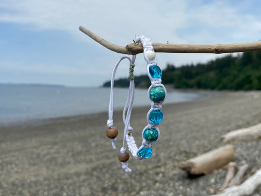 A beautiful white woven bracelet with ocean tome blue green beads, and a white lava bead, with wood beads dangling from the adjustment cords hanging on a piece of driftwood.