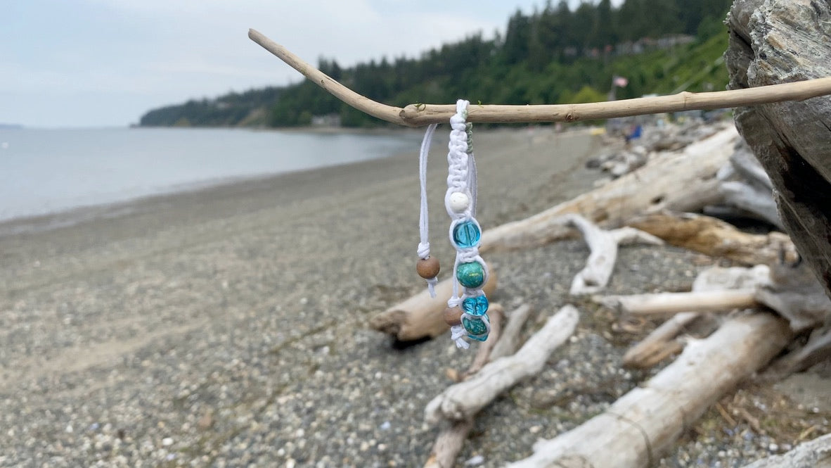 A beautiful white woven bracelet with ocean tome blue green beads, and a white lava bead, with wood beads dangling from the adjustment cords hanging on a piece of driftwood.