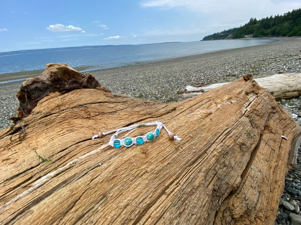 A beautiful white woven bracelet with ocean tome blue green beads, and a white lava bead, with wood beads dangling from the adjustment cords sitting on a piece of driftwood.