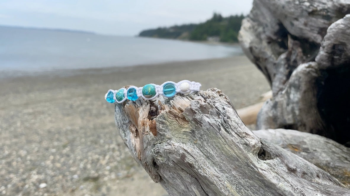 A beautiful white woven bracelet with ocean tome blue green beads, and a white lava bead, with wood beads dangling from the adjustment cords sitting on a piece of driftwood.
