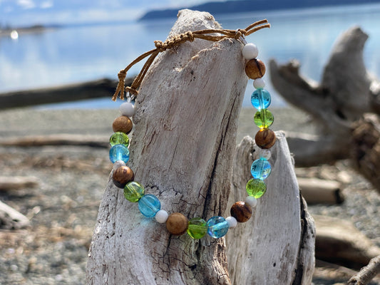 Beautiful beach vibe bracelet with green and blue  ocean colored beads with small white turqoise beads, and wood grain beads, with adjustable strap on a piece of driftwood with the ocean in the background
