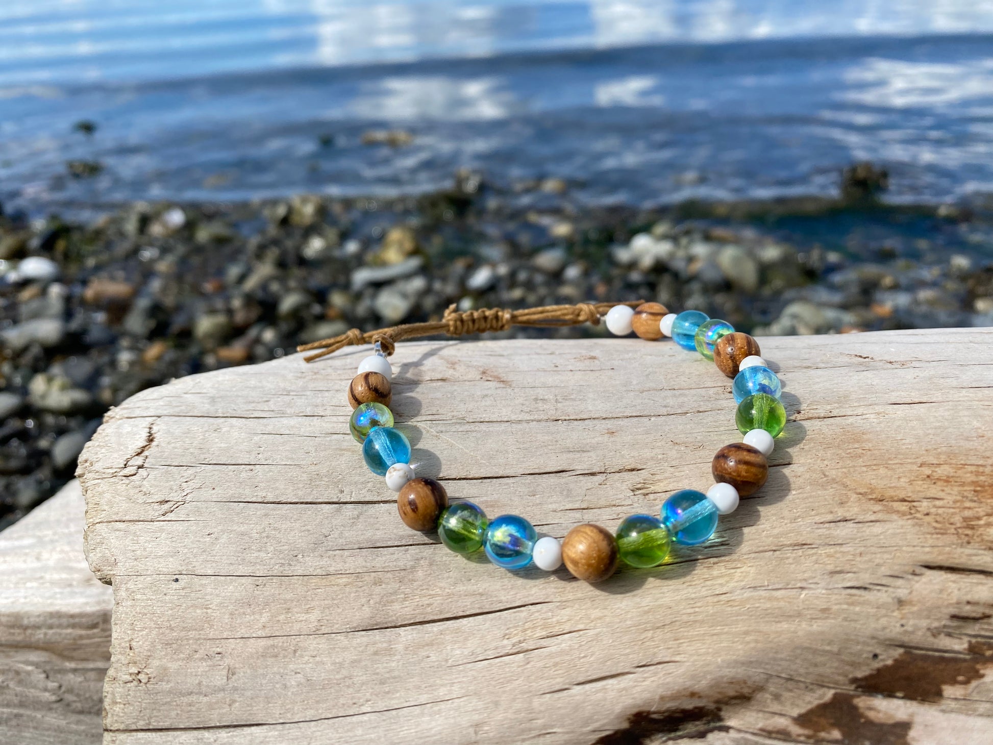 Beautiful beach vibe bracelet with green and blue  ocean colored beads with small white turqoise beads, and wood grain beads, with adjustable strap on a piece of driftwood with the ocean in the background