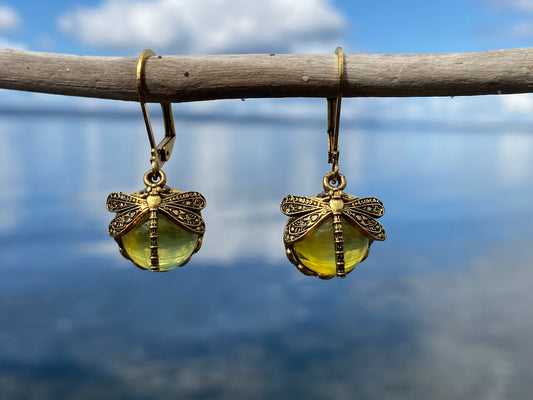 Beautiful dragonfly earrings with yellow sparkling glass and antique gold dragonflies over the top, hanging on a stick with the ocean behind
